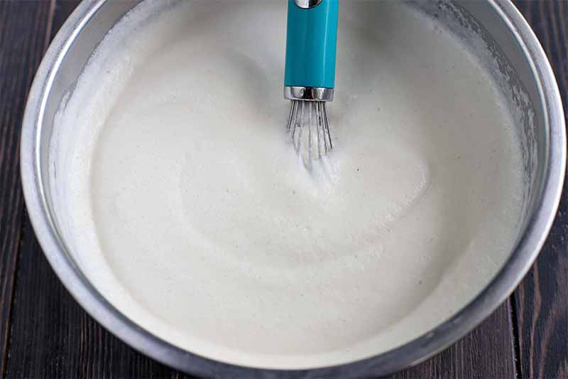Soft whipped egg whites in a stainless steel mixing bowl, with a blue and silver wire whisk, on a brown wood background.