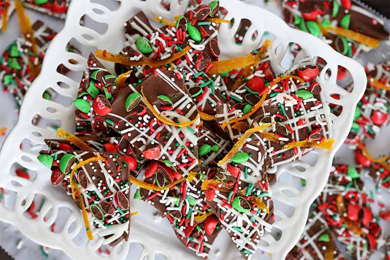 A white decorative serving plate of homemade candy bark for the holidays, with more scattered on a table in the background.
