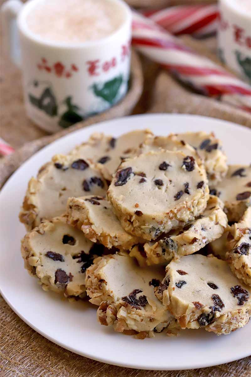A white plate of slice and bake butter cookies studded with raisins, with a mug of eggnog and candy canes in the background, on light brown burlap.