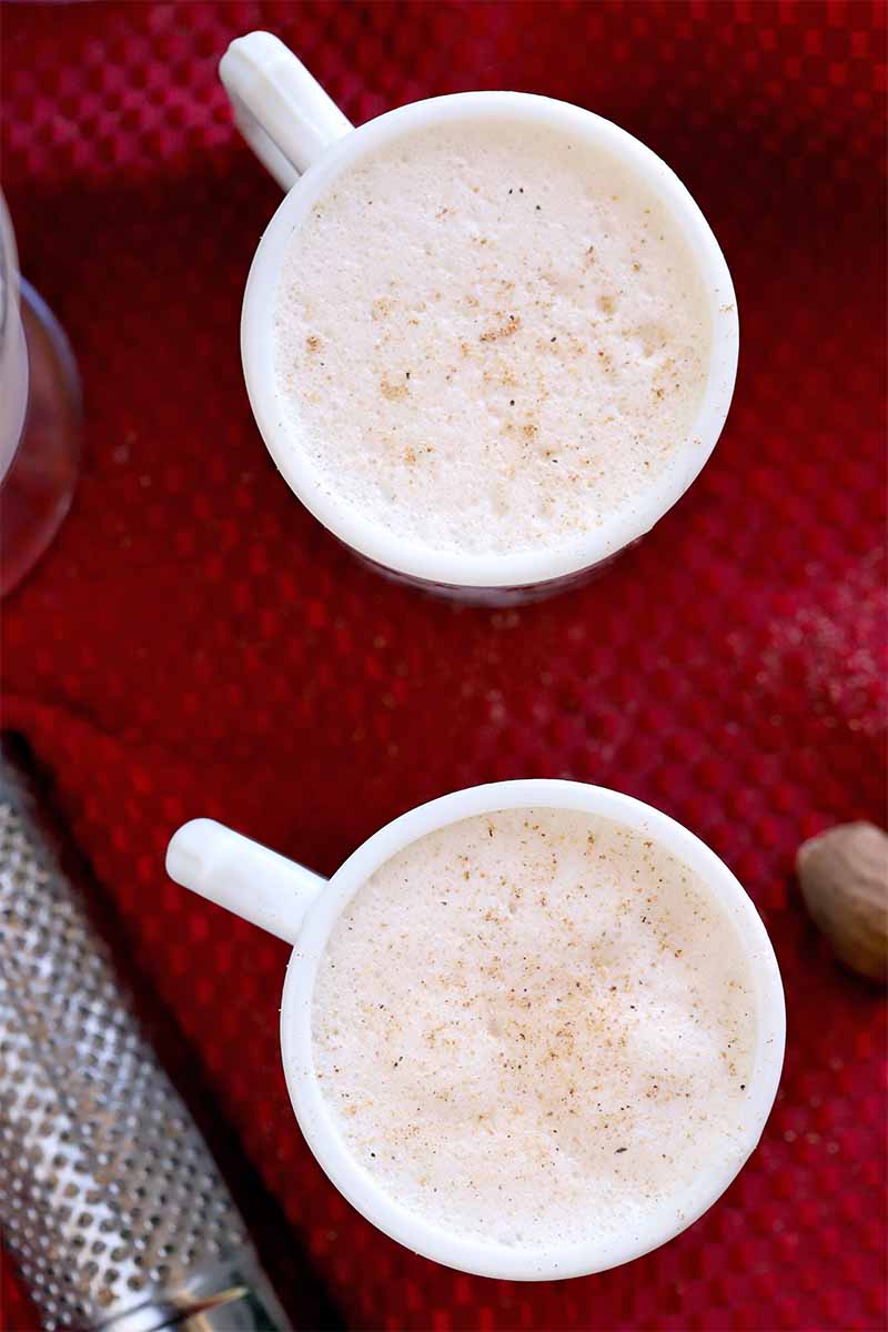Overhead shot of two white mugs filled with eggnog and topped with a sprinkling of nutmeg, on a red kitchen towel with the whole spice and a metal grater.