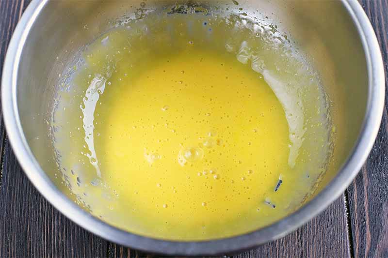 Whipped egg yolks in the bottom of a stainless steel mixing bowl, on a dark brown wood table.