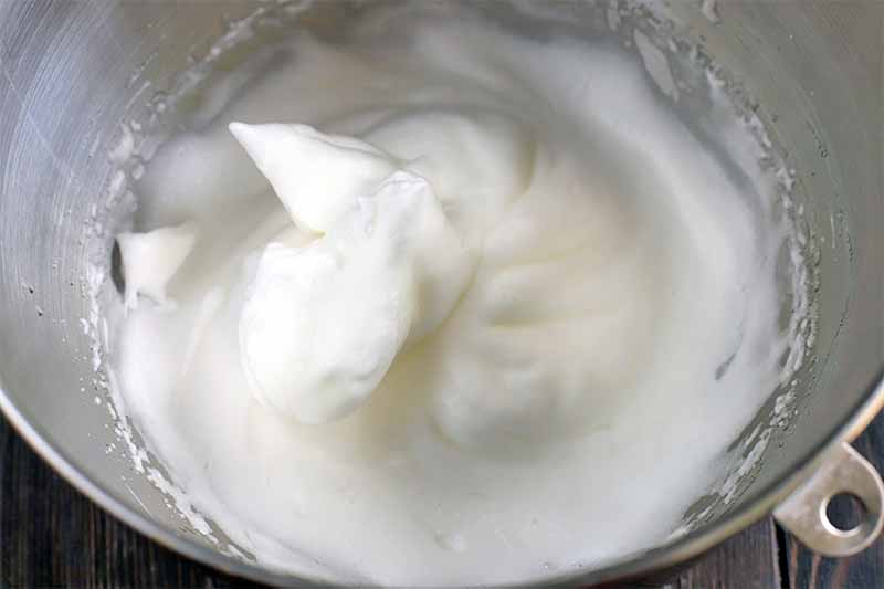 Egg whites whipped to form soft peaks are in a large stainless steel stand mixer bowl, on a dark brown wood surface.