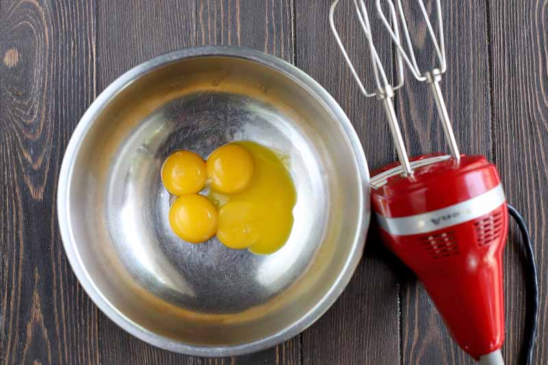 Four egg yolks are in a stainless steel bowl to the left of a red KitchenAid hand mixer with silver beaters, on a dark brown wood surface.