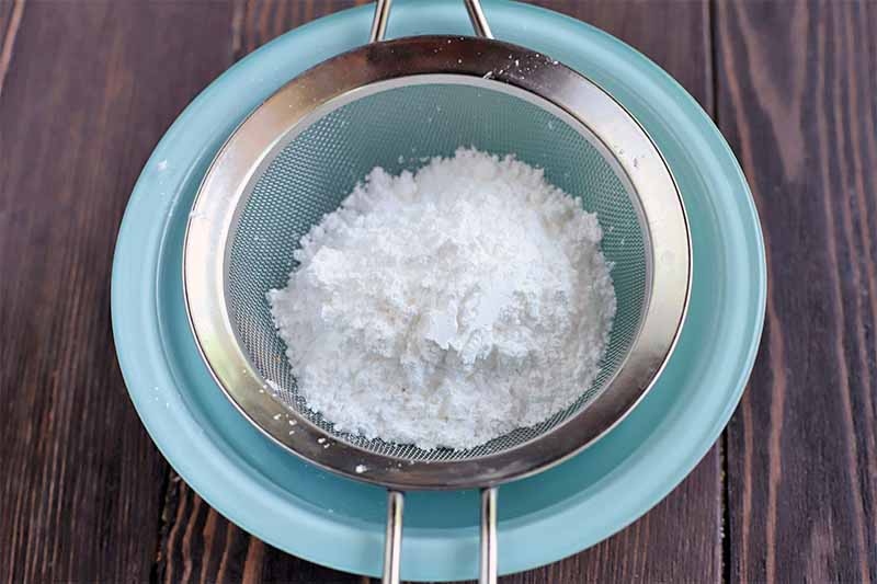 Confectioners' sugar is being sifted through a fine mesh metal sieve into a light blue glass bowl, on a brown wood surface.