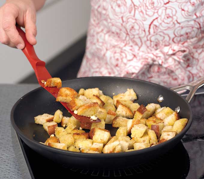 Closeup of the torso of a person clothed in a white and red floral blouse, holding a red plastic spatula and flipping over piece of bread that are tosating in a frying pan to make croutons, on an electric hotplate.