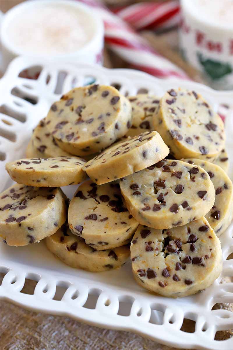 Round butter cookies studded with mini chocolate chips are arranged in a decorative white ceramic serving dish, with white mugs of eggnog and red and white striped candy canes in the background.