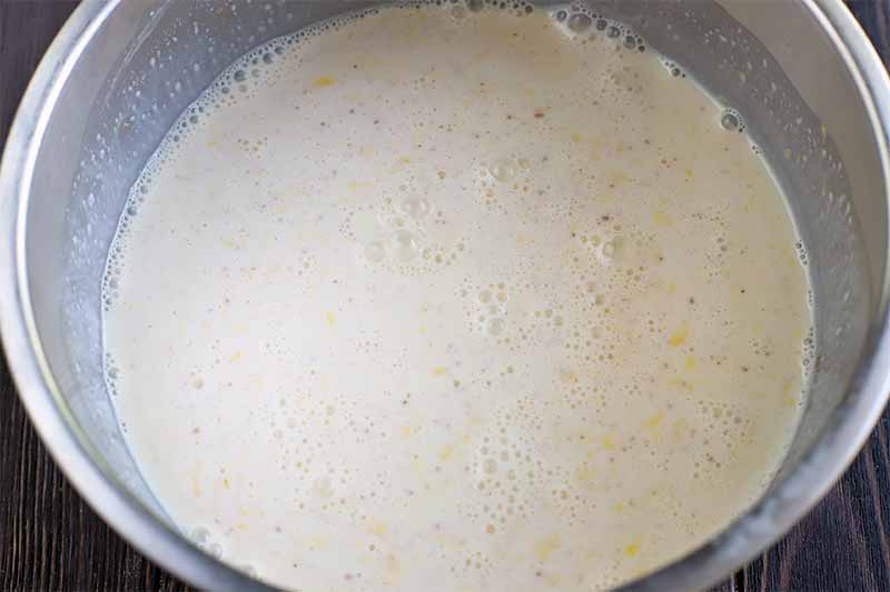 A frothy egg and cream mixture is in a large stainless steel mixing bowl, on a dark brown wood surface.