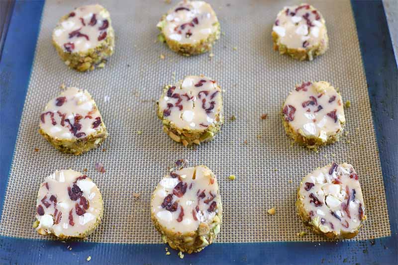 Cranberry and white chocolate cookie dough sliced into round portions and rolled in crushed pistachios, arranged in three rows of three on a blue and tan silicone baking pan liner.