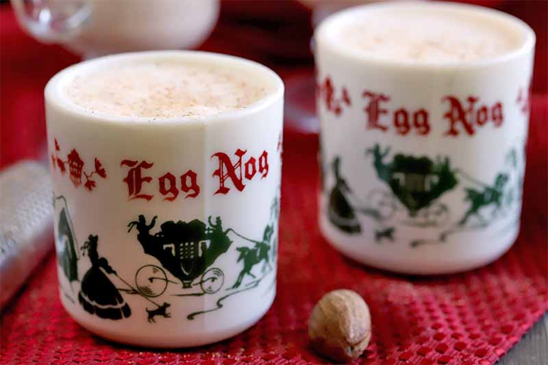 Two decorative red, green, and white holiday-themed mugs of eggnog, with a whole nutmeg and metal grater on a red kitchen towel that is spread over a dark brown wood table.