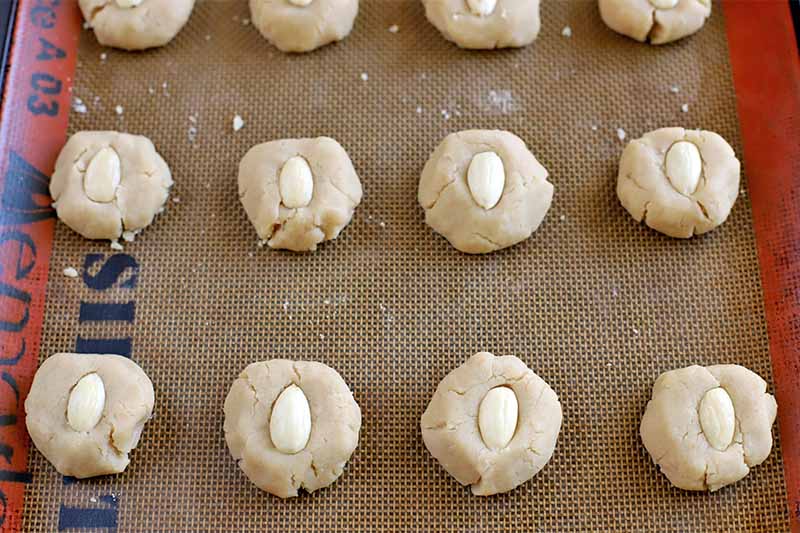 Flattened balls of cookie dough with half of a blanched almond pressed into the top of each are arranged in three rows on a silicone pan liner.