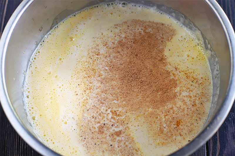 A homemade eggnog mixture in a larger stainless steel bowl with ground nutmeg sprinkled on the surface, on a dark brown wood background.