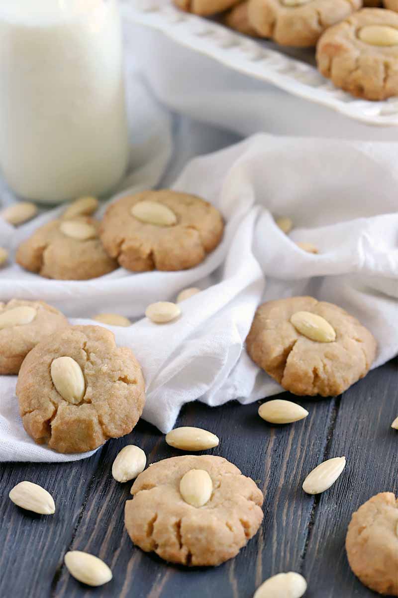 Brown butter cookies on a gathered white cloth, with scattered blanched almonds on a bronw wood surface, with a glass bottle of milk and a white ceramic serving dish in the background.
