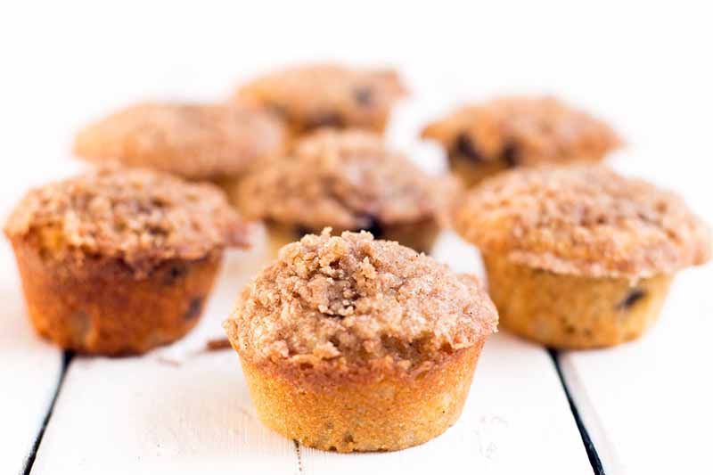 Oblique shot of seven blueberry muffins with crumble topping shot with selective focus, on a white wood background.