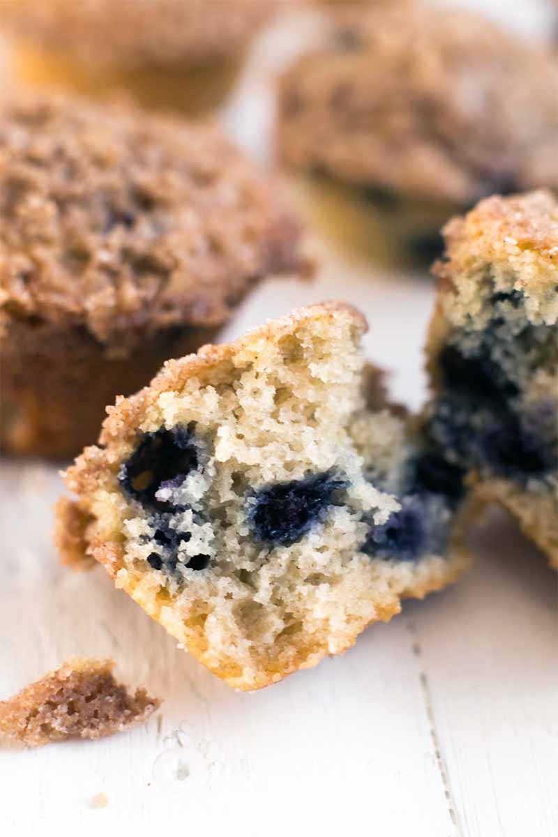 Vertical image of a homemade vegan blueberry muffin with crumble topping, torn in half to show the crumb inside, on a white painted wood surface with more of the baked goods in soft focus in the background.