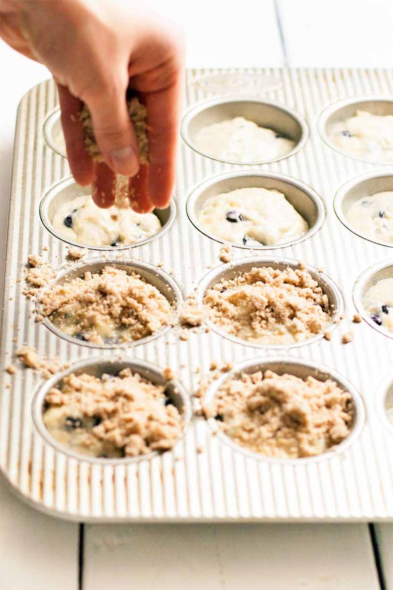 A hand crumbles streusel topping onto muffin batter in a baking pan, on a white painted wood surface.