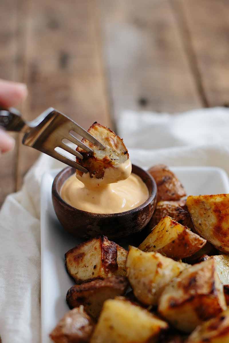 Vertical image of a fork piercing a roasted potato dipped in a mayo sauce on a plate with more roasted potatoes.