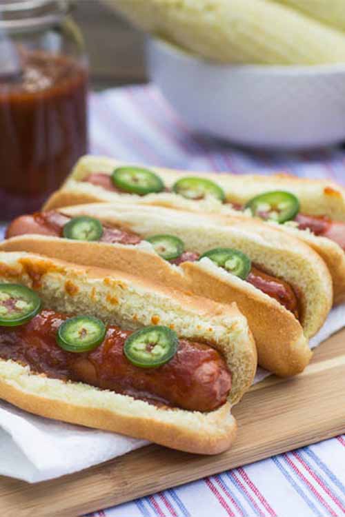 Three hot dogs topped with barbecue sauce and sliced jalapenos, on a wood board topped with a paper towel, on a red, white, and blue striped cloth kitchen towel, with a bowl of cooked ears of corn in the background.