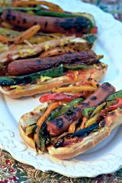 Grilled frankfurters in buns with bell peppers, on a white serving platter.