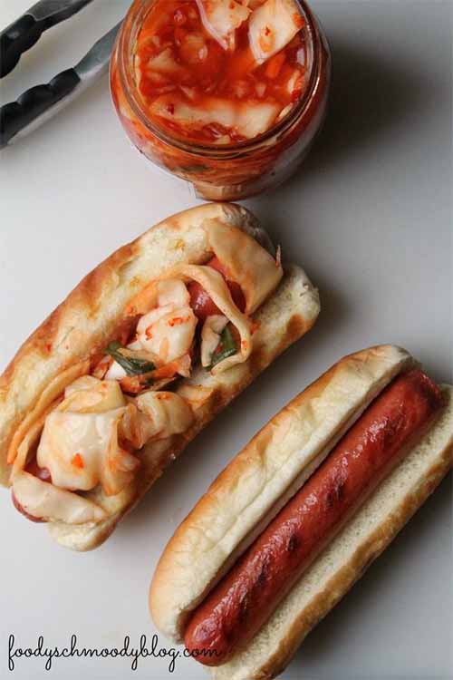 Two grilled franks in buns, with the one on the left topped with kimchi and a jar of the same in the background next to a pair of tongs, on a white surface.