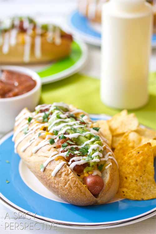 Vertical head-on image of a hot dog topped with green sauce and a drizzle of white sauce, on a blue and white plate with a small pile of corn chips, with more plated sandwiches, a squeeze bottle of sauce, and a white bowl of ketchup in the background.