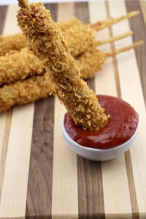 Three deep-fried Pringles-coated hot dogs on sticks rest on a striped wooden serving board in the background, with another being dipped into a small cup of ketchup in the foreground.