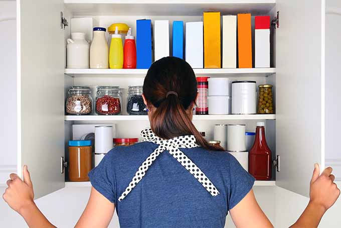 A woman with brown hair pulled back into a ponytail, wearing a blue t-shirt and a black and white checked apron, peers into the cupboard while the holds the doors open.