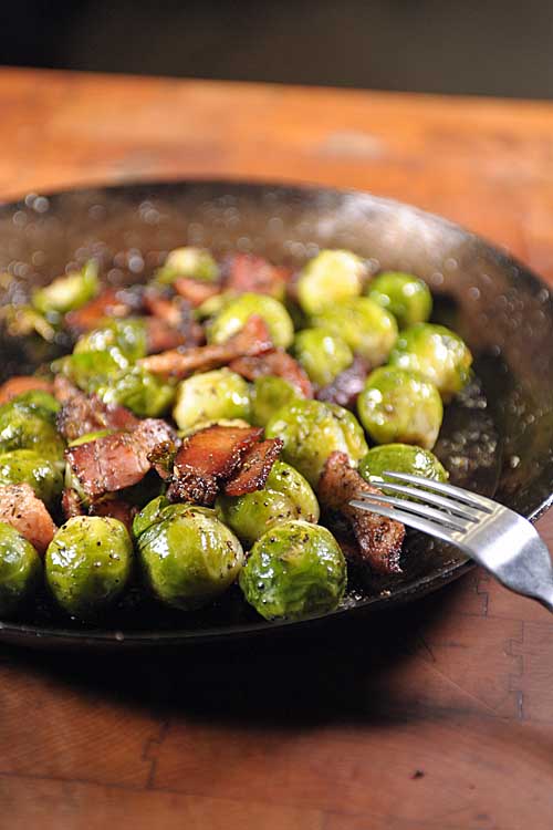 Closely cropped vertical image of a fork resting on the side of a well-worn carbon steel pan of whole Brussels sprouts with bacon, on a wood background.