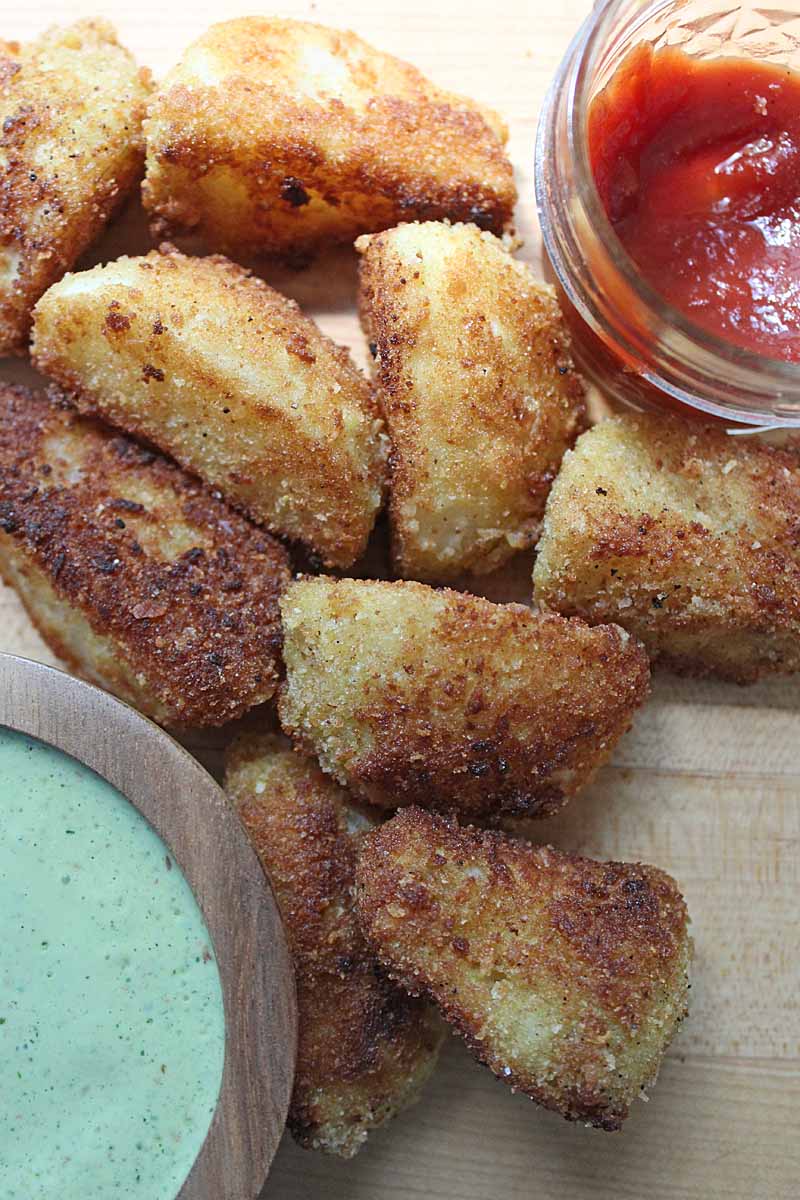 Top down view of a batch of fried kohlrabi wedges with a container of cilantro yogurt dipping sauce and a second container of ketchup.