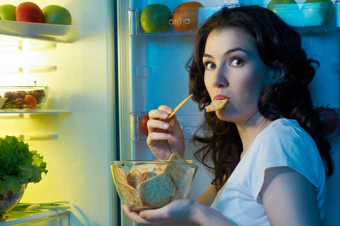 An attractive lady is caught late at night with the refrigerator door open and eating food with a guilty look on her face. | Foodal.com