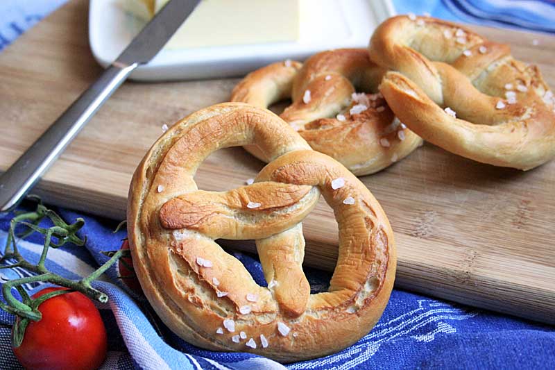 A close up of a traditional German lye pretzel centered in the frame with a cutting board and a stick of butter in the background. A red cherry tomato sits to the lower left.