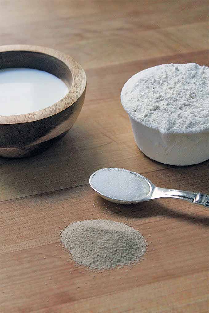 Sugar, flour, and yeast in measuring cups and a spoon on a wooden surface.
