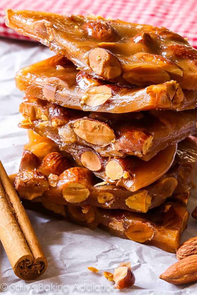 Vertical image of a stack of pieces of almond toffee, with whole nuts and a cinnamon stick on a crumpled piece of parchment paper, with a red and white checkered cloth in soft focus in the background.