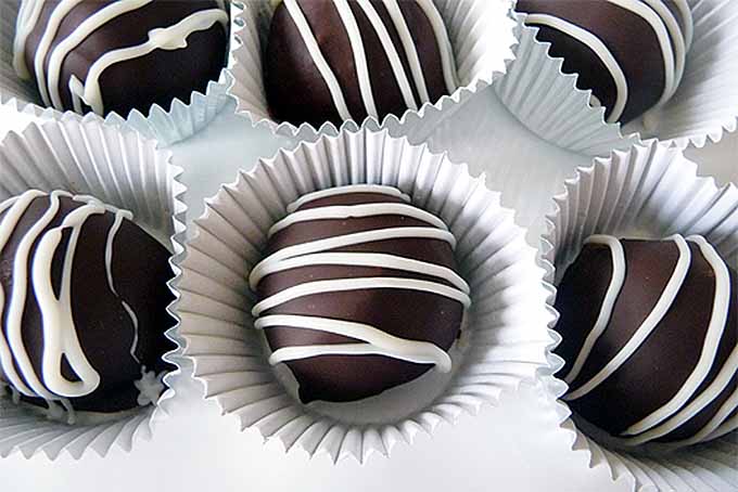 Horizontal overhead closely cropped image of six dark chocolate truffles drizzles it a decorative white zig-zag, in white paper wrappers on a white surface.