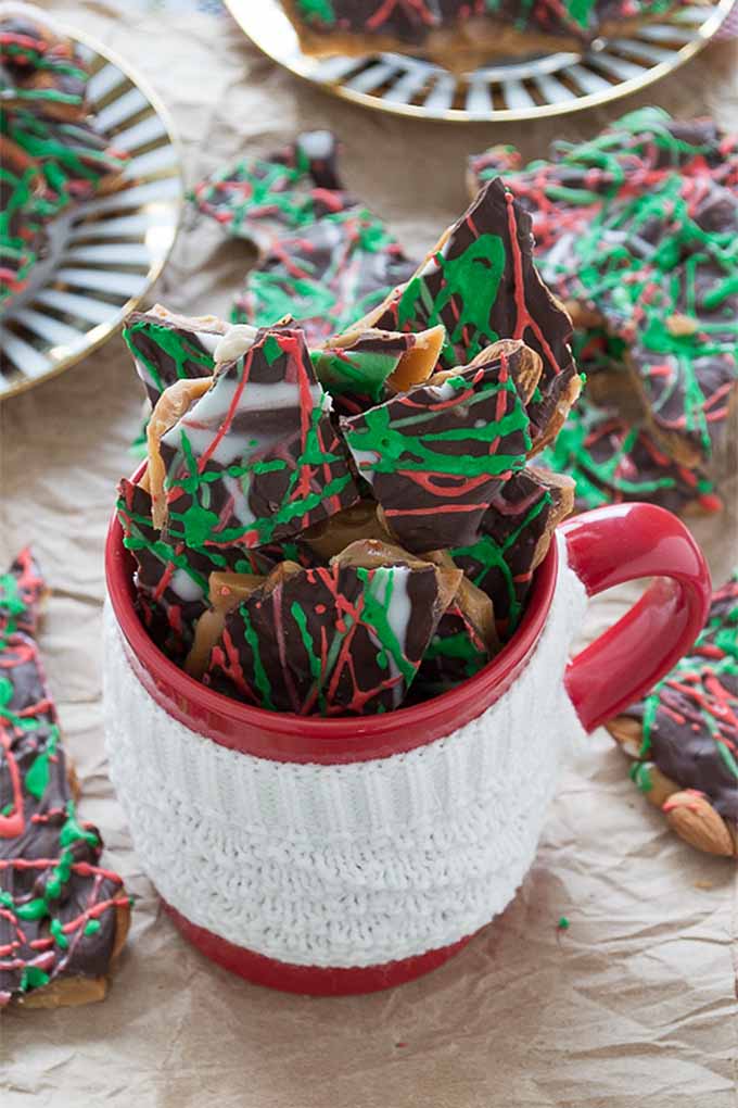 Vertical oblique overhead image of a red mug with a white crocheted hand protector, filled with chocolate toffee decorated with stripes of red, white, and green, on a crumpled piece of brown paper with more pieces of the treats on gold and white plates and scattered around the mug.