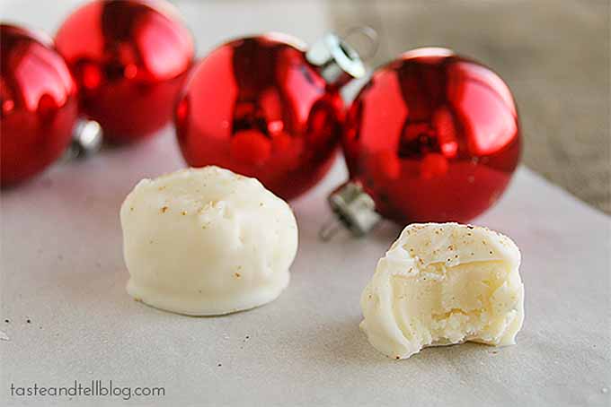 Two white eggnog truffles sprinkled with nutmeg, one with a bite taken out of it to show the inside, on a gray surface with four red glass Christmas ball ornaments in soft focus in the background.