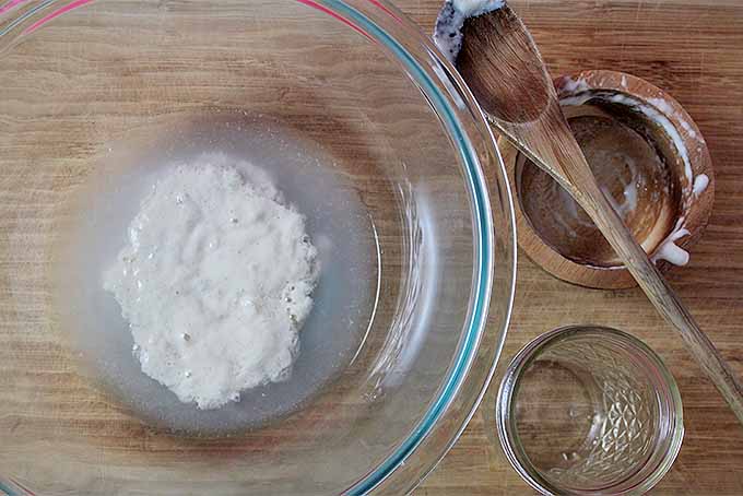 Top view of sourdough starter in a glass mixing bowl, with a wooden spoon, ramekin, and drinking glass to the right of the bowl on a wooden table.