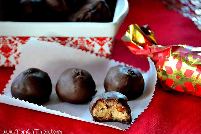 Horizontal image of four homemade maple nut chocolates on a white piece of parchment paper with a crimped edge, on a red table cloth with a tray of more candy in the background, and a gold and red paper-wrapped Christmas cracker to the right.