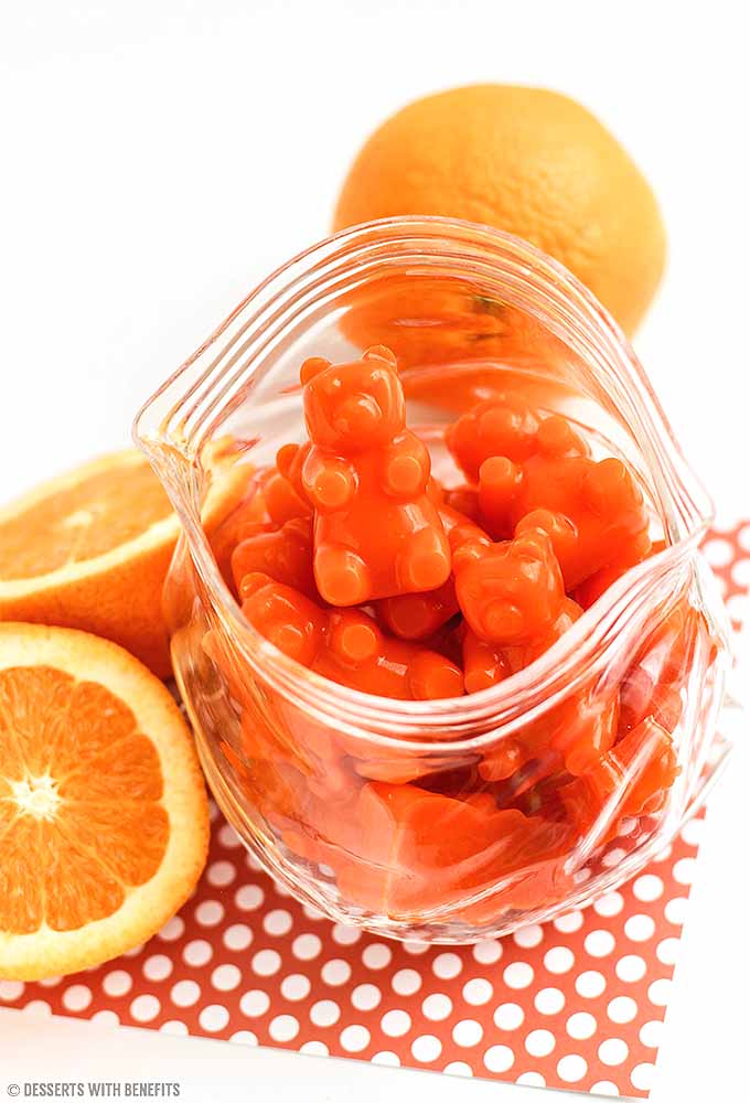 Vertical image of homemade orange gummy bears in a clear glass dish in the shape of a zip-top plastic bad, on an orange cloth with white dots, with a whole and sliced orange on a white background.