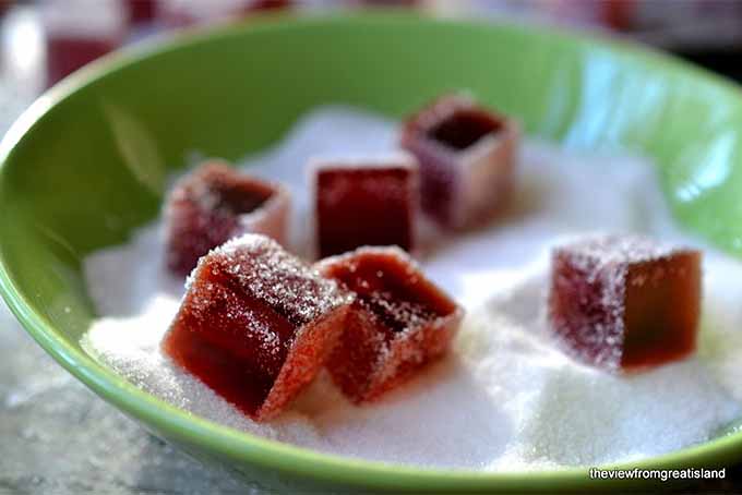 Square homemade pomegranate gumdrops in a green shallow ceramic dish of granulated sugar for coating.