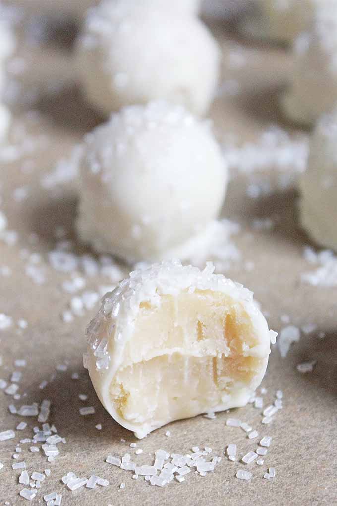 Vertical image of sugar cookie truffles decorated with large white sugar crystals, with a bite taken out of the one in the front to show the inside, on a gray countertop.