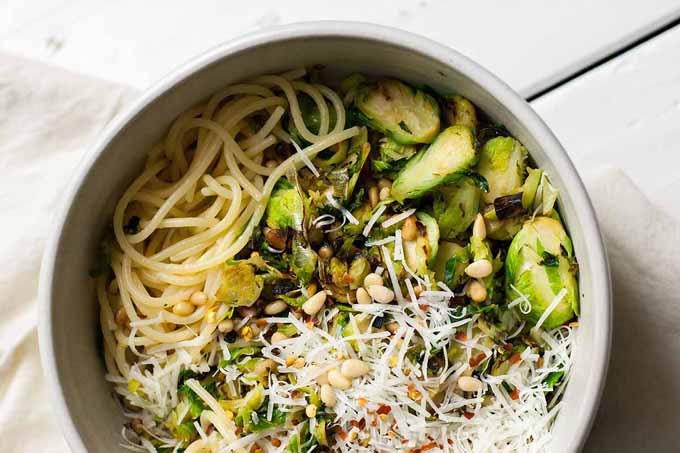 Top down view of bowl full of spaghetti noddles topped with shaved Brussels sprouts, leeks and pine nuts.