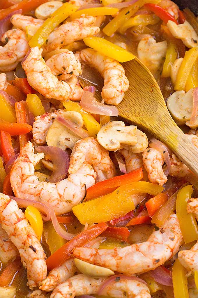Closeup overhead shot of cooked shrimp and stripe of red and yellow bell pepper being stirred with a wooden spoon.