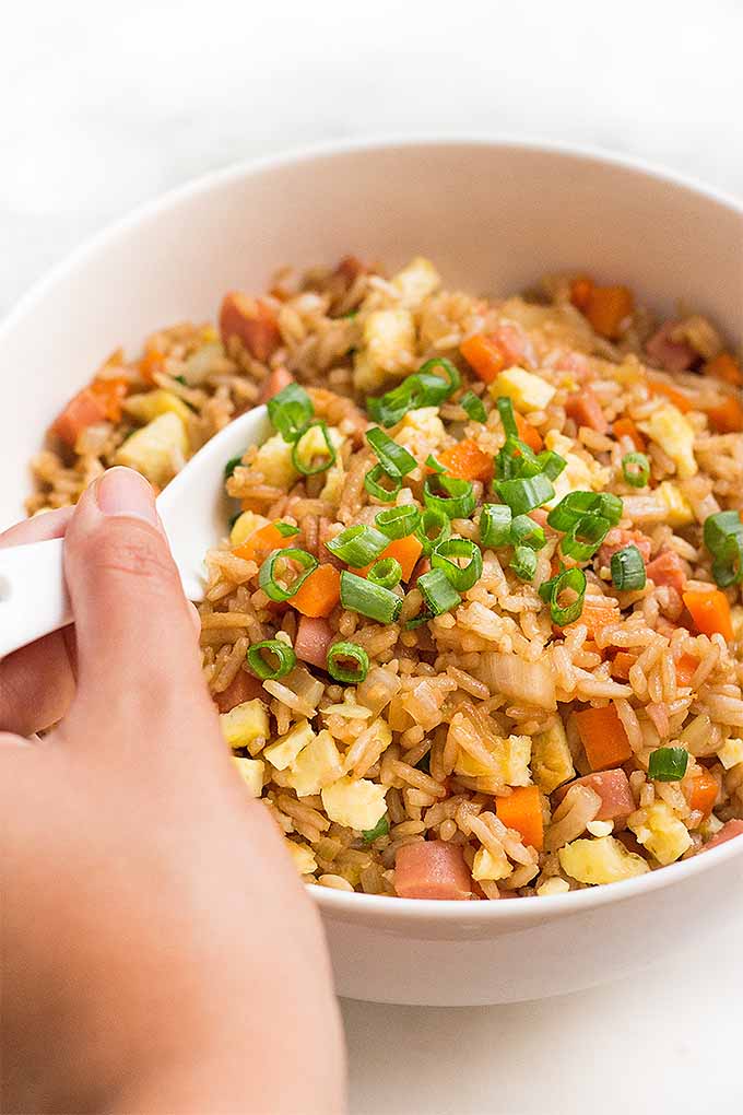 Fried rice with hot dogs, carrots, onion, ginger, garlic, and chopped green scallions, in a white bowl with a hand holding a ceramic spoon.