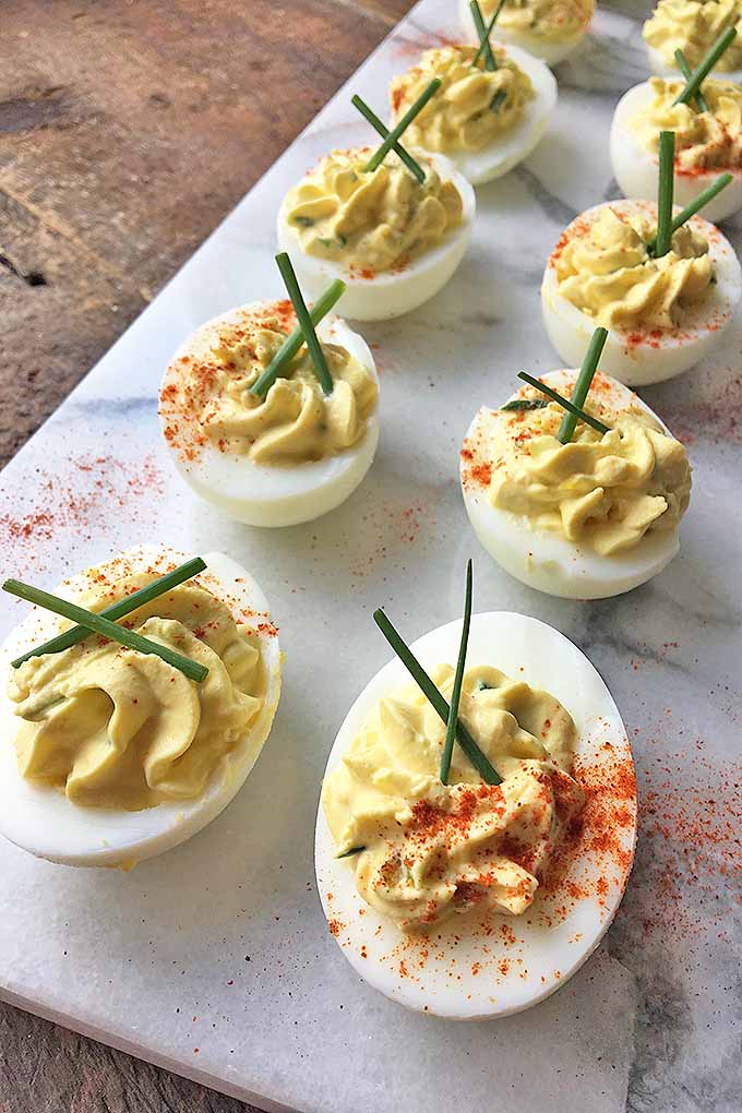 Vertical image of of deviled eggs with paprika and chives on a marble cutting board on a wooden surface.