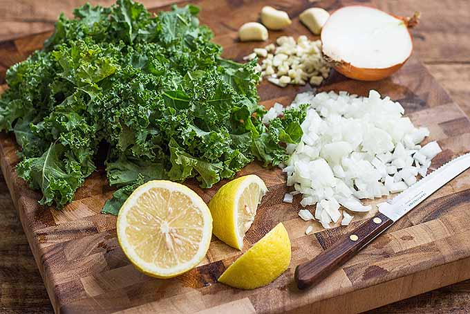 Chopping up onions, kale, and garlic and slicing up lemon on a wooden cutting board.