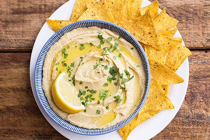 Overhead image of a blue and white bowlful of baba ghanoush hummus with a lemon wedge, olive oil, and herbs, on a white plate with yellow corn chips, on a brown wooden background.