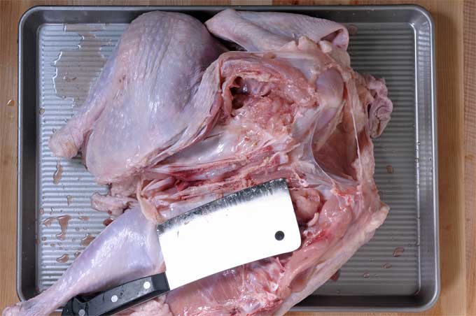 A turkey whose breast bone has been cracked with a meat cleaver. Laying in a baking tray with the meat cleaver resting on top of it. | Foodal
