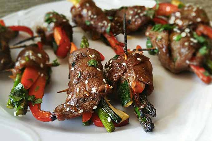 Asparagus, bell pepper, and steak roll-ups with herbs and sesame seeds on a white plate.