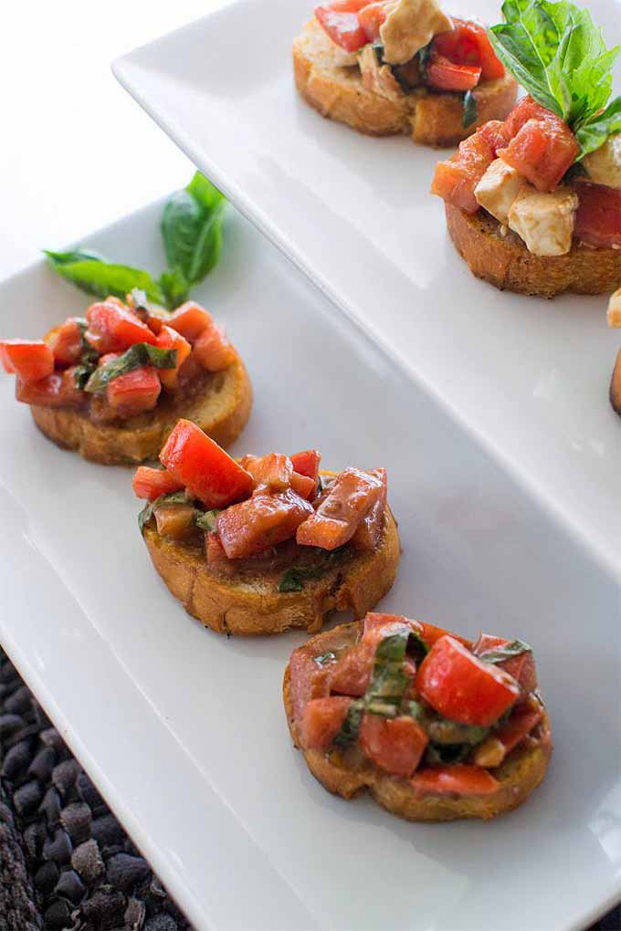 Bruschetta with mozzarella, tomato and basil on toast rounds, on white stacking serving trays with sprigs of fresh herbs.