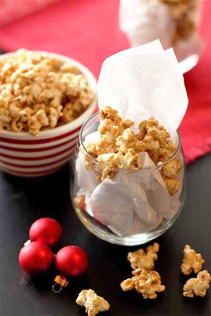 A bowl and paper-filled glass of caramel popcorn, on a black table with red sphere-shaped Christmas ornaments and a red placemat.